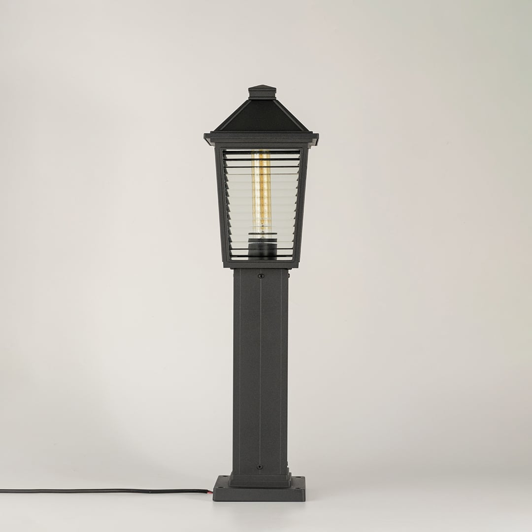 A black outdoor pillar lamp with ribbed glass shown with light on against a white background highlighting its warm glow and elegant tapered lantern design.