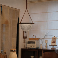 A rustic pendant light with a textured, hammered silver dome shade suspended by brown leather straps from a dark wooden rod, hanging in a cozy, vintage-style room with a wooden cabinet and neutral-toned decor.