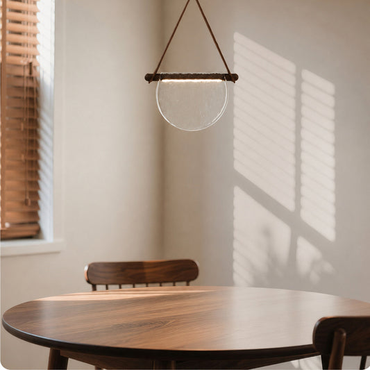 A modern pendant light with a clear glass bowl and a dark brown wooden top bar hangs over a round wooden dining table in a softly lit room with window blinds casting shadows on the wall.