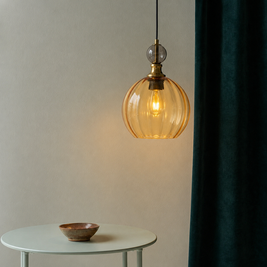 A hanging pendant light with a ribbed amber glass shade and brass fixture, shown lit in a styled interior scene with a small round table and a dark teal curtain.