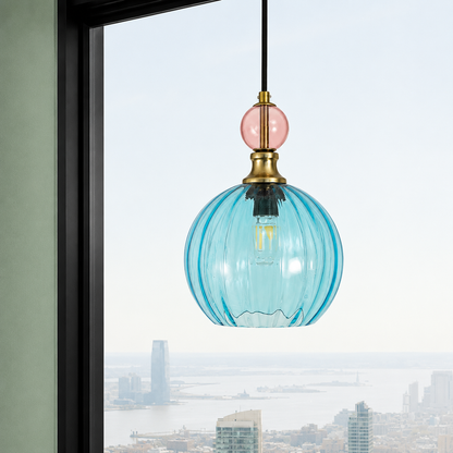 A modern pendant light with a ribbed, translucent light blue glass shade, a round pink glass accent, and a brass metal cap, hanging in front of a window with a city skyline view.