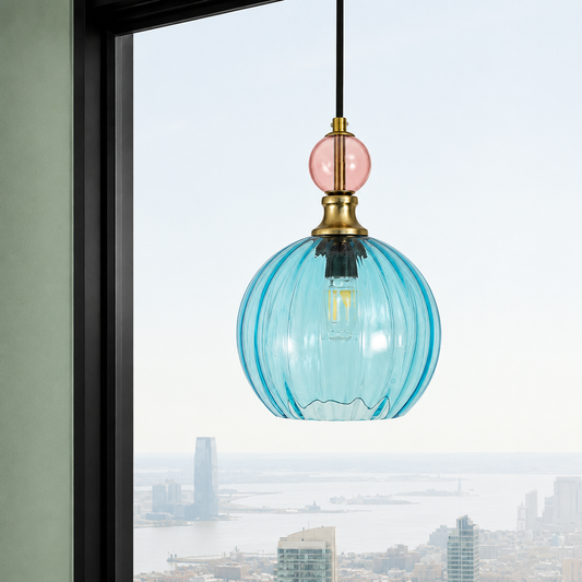 A modern pendant light with a ribbed, translucent light blue glass shade, a round pink glass accent, and a brass metal cap, hanging in front of a window with a city skyline view.