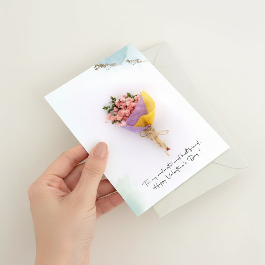 Hand holding a white greeting card featuring a mini pink and yellow dried flower bouquet over a clean minimalist background.