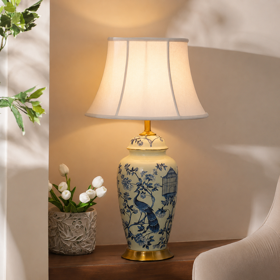 A ceramic table lamp with a blue and white peacock pattern on a beige vase-style base, topped with a gold finial and a white pleated drum shade. The lamp is lit and placed on a wooden table in a warm indoor setting next to a small vase of white tulips.