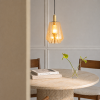 A modern pendant light with a warm glowing glass shade and brass-toned metal accents hangs above a round dining table in a softly lit interior with wooden chairs and a small vase.