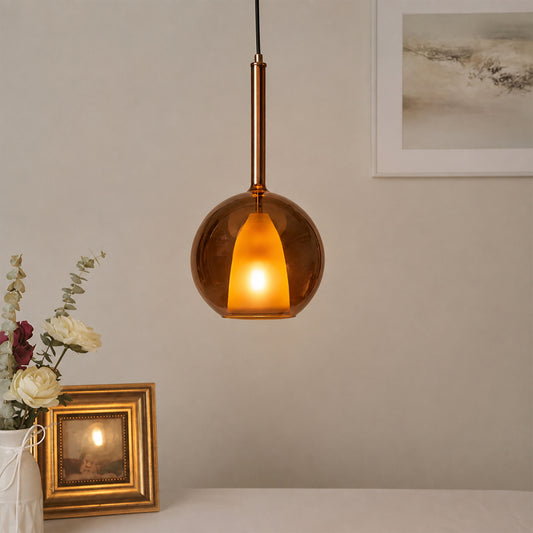 A modern pendant light with a spherical amber-brown glass shade and a warm glowing light inside, hanging in a styled interior setting with a vase of flowers and a picture frame below.
