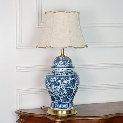 Classic blue white ceramic table lamp with ornate floral design brass base and fabric shade on off white background.