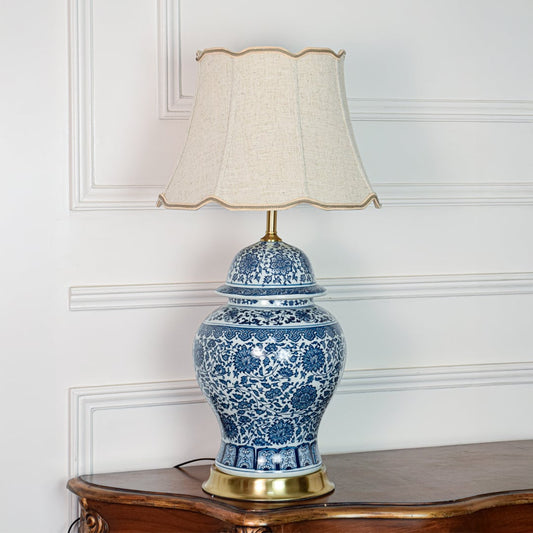 Classic blue white ceramic table lamp with ornate floral design brass base and fabric shade on off white background.