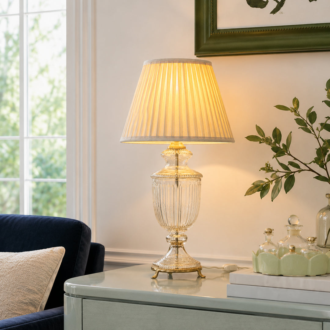 Crystal glass table lamp with pleated shade glowing warmly on side table beside window in elegant living room.