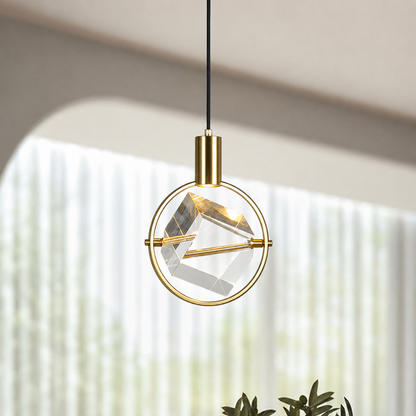 A modern gold pendant light with a circular brass frame and a faceted clear glass geometric shade, suspended by a black cord in a softly lit interior setting.
