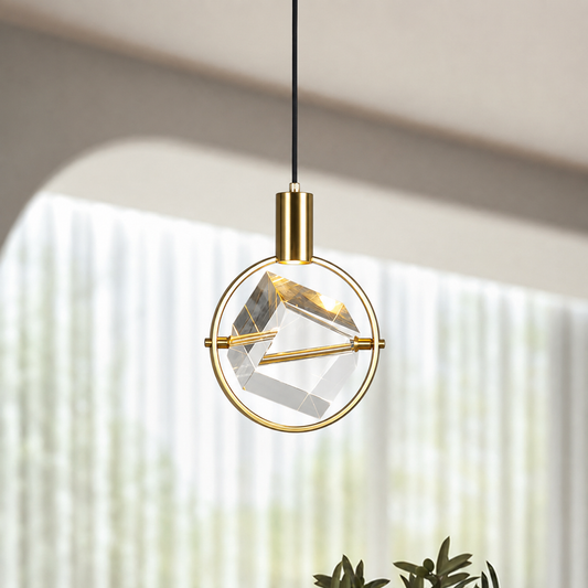 A modern gold pendant light with a circular brass frame and a faceted clear glass geometric shade, suspended by a black cord in a softly lit interior setting.