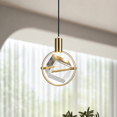 A modern gold pendant light with a circular brass frame and a faceted clear glass geometric shade, suspended by a black cord in a softly lit interior setting.
