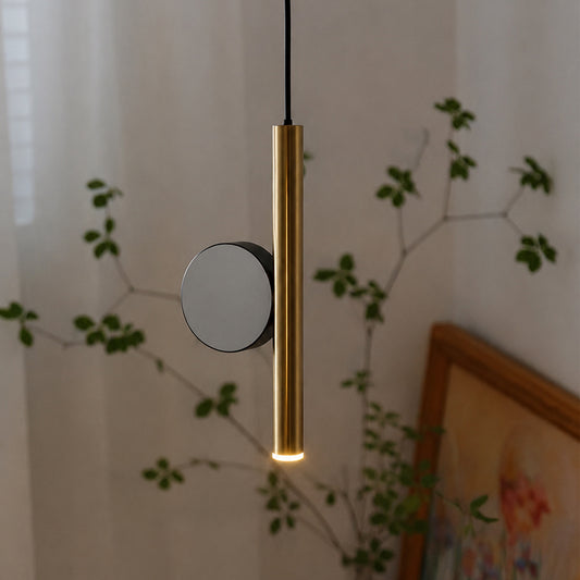 A modern cylindrical pendant light with a brushed gold finish and a circular black accent, shown hanging in a softly lit room with blurred greenery and a framed picture in the background.