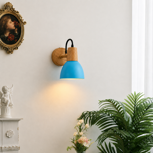 A modern wall sconce light fixture with a bright blue dome shade, natural wood accents, and a black cord, mounted on a white wall in a styled interior setting with a plant and framed art.