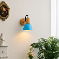 A modern wall sconce light fixture with a bright blue dome shade, natural wood accents, and a black cord, mounted on a white wall in a styled interior setting with a plant and framed art.