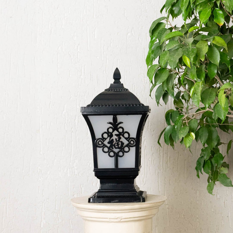 Black outdoor pillar light with ribbed glass Light On placed next to a lush green plant Lifestyle image for modern patio decor.