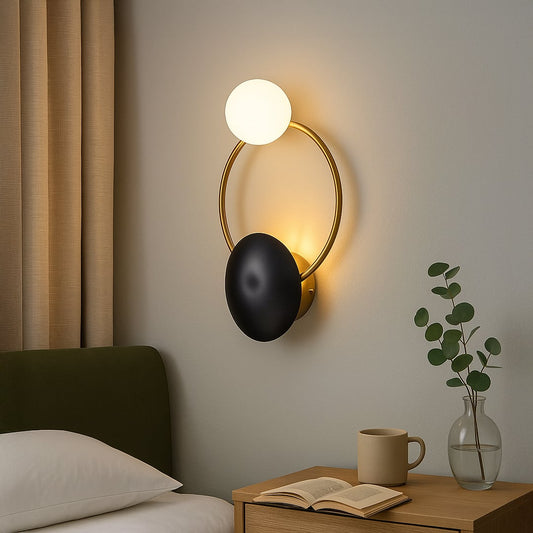 Modern gold ring wall light with black disc and white glass globe mounted next to bed for contemporary bedroom reading lighting.