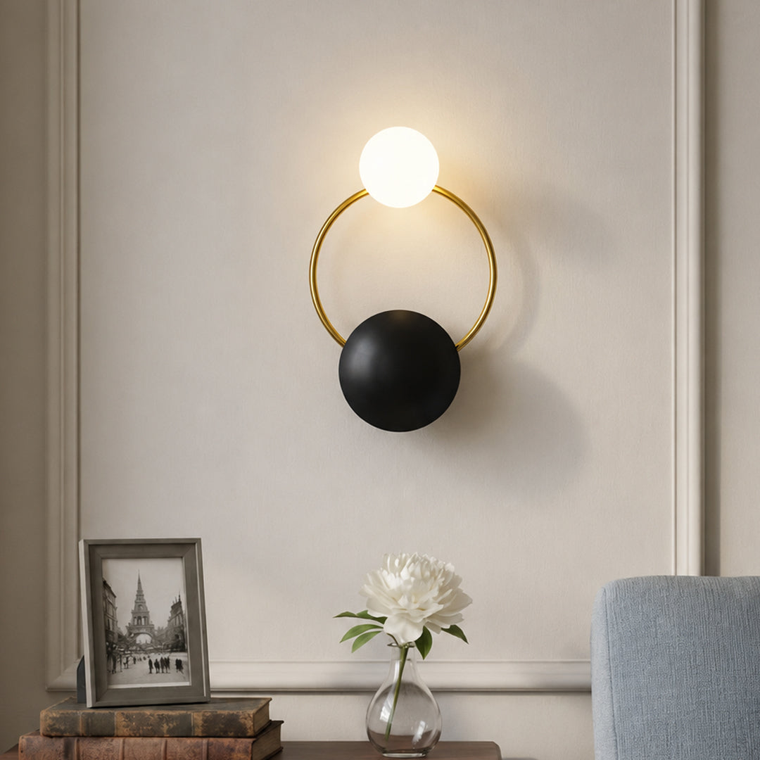 Modern gold brass hoop wall sconce with black plate accent and glowing glass orb bulb mounted in a luxury living room.