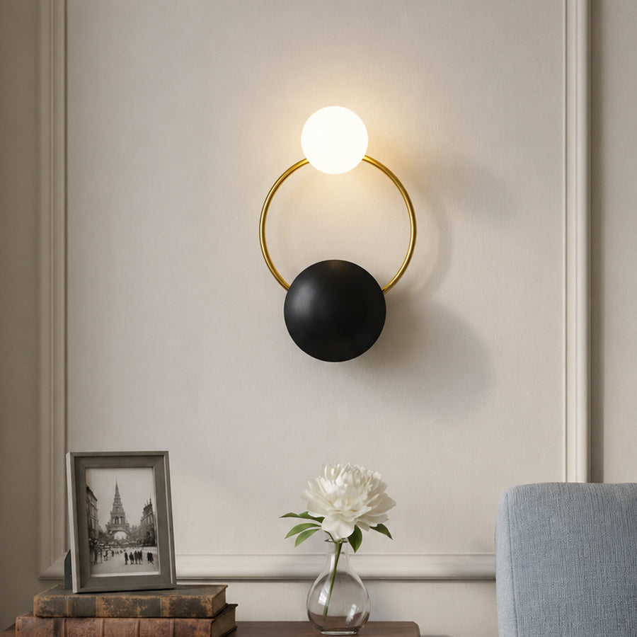 Modern gold brass hoop wall sconce with black plate accent and glowing glass orb bulb mounted in a luxury living room.