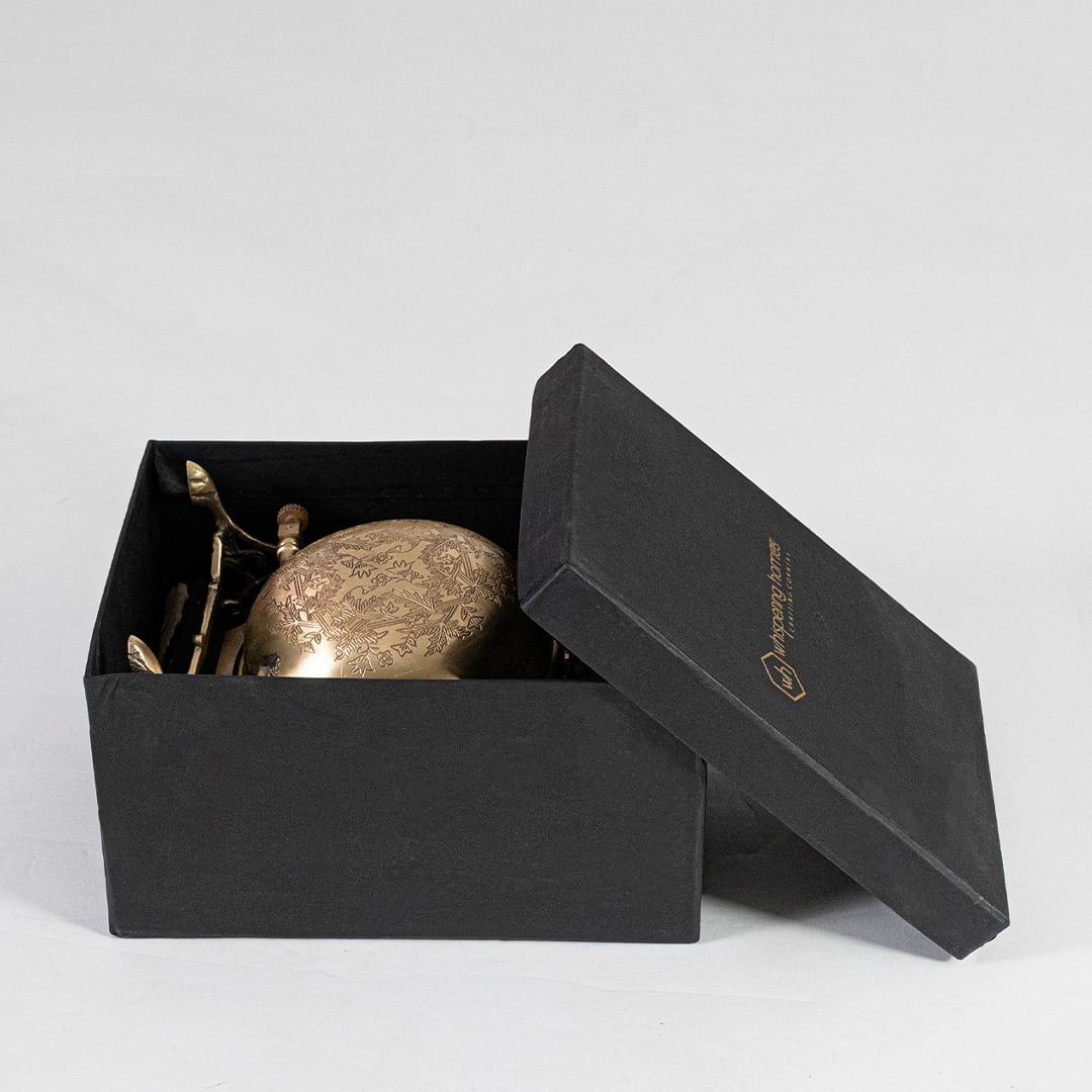 A brass swing bowl in box with detailed engraving is shown against a white background focusing on the reflective metal surface and the intricate handcrafted stand.