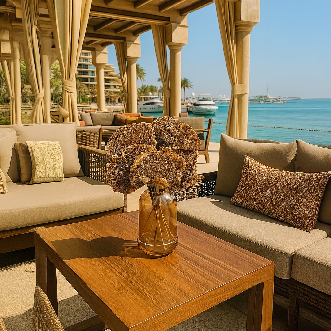 Dried brown fan stems arranged in a glass vase on a wooden outdoor table styled with beige seating and oceanfront decor in natural light.