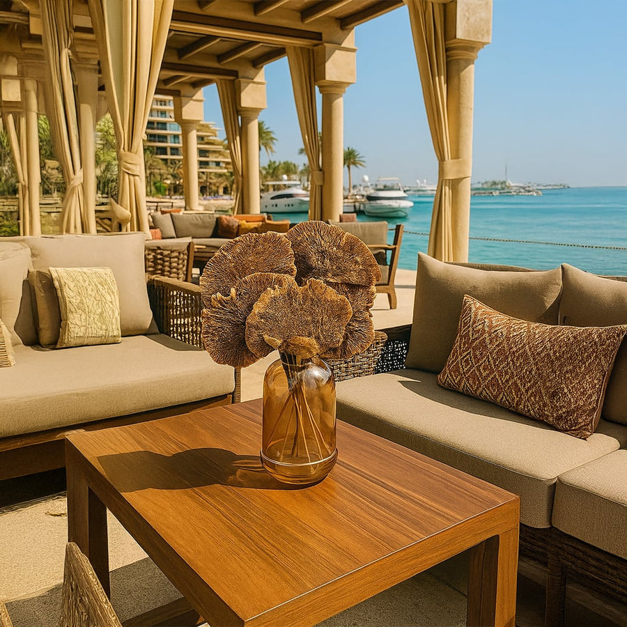 Dried brown fan stems arranged in a glass vase on a wooden outdoor table styled with beige seating and oceanfront decor in natural light.