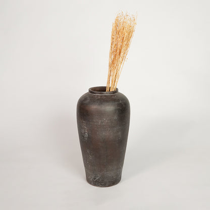 Natural beige dried decorative floral stems arranged inside a tall dark brown rustic ceramic vase on white backdrop.