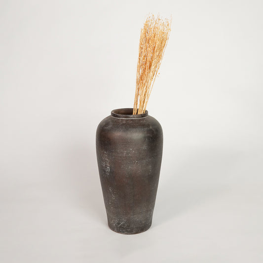 Natural beige dried decorative floral stems arranged inside a tall dark brown rustic ceramic vase on white backdrop.