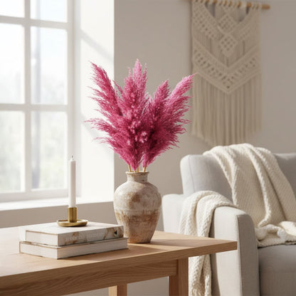 Neutral ceramic vase with pink dried pampas stems styled on a wooden table beside an armchair in a soft modern living room interior setting.
