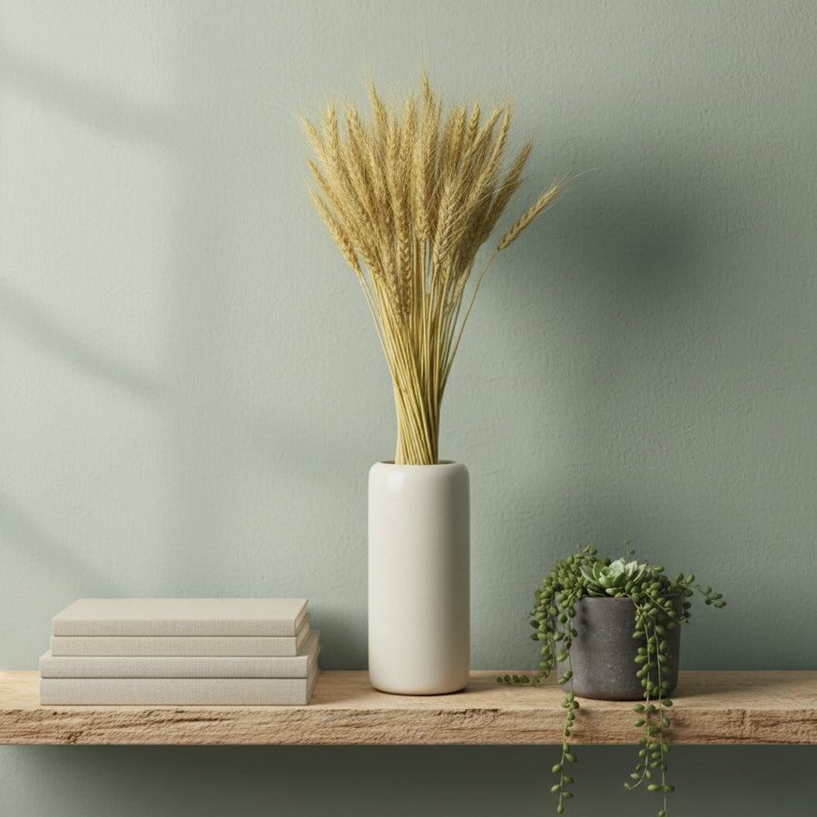 Natural golden dried wheat stalks in a white cylindrical vase on a rustic wooden shelf against a sage green wall.