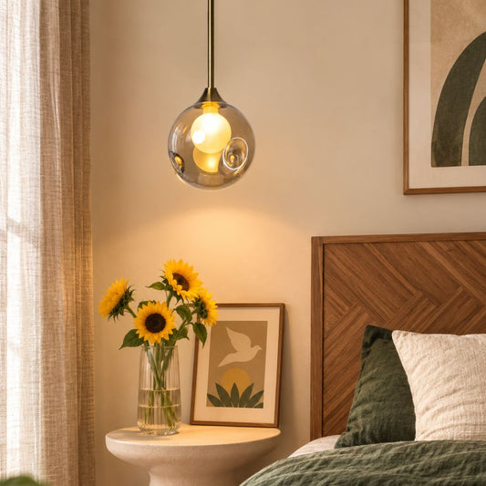 A modern pendant light with a clear glass globe shade and a glowing warm bulb, hanging above a bedside table in a softly lit bedroom. Sunflowers in a vase, a wooden headboard, and framed wall art are visible in the room.