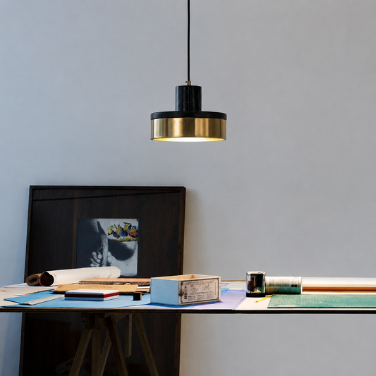 A modern pendant light with a black top and a brushed gold lower band hangs over a desk in a minimalist room setting.