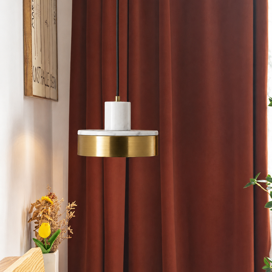 A modern pendant light with a white marble cylinder and a brushed gold metal band, hanging in front of rust-brown curtains in a styled interior setting.
