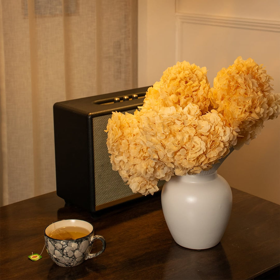 Yellow preserved hydrangea flowers in a ceramic vase on a wooden side table next to a retro speaker and coffee cup.