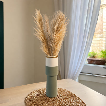 A modern ceramic vase with a matte sage green and white color-blocked design, topped with a gold band and filled with dried pampas grass, displayed on a woven placemat on a light wood table in a bright room.