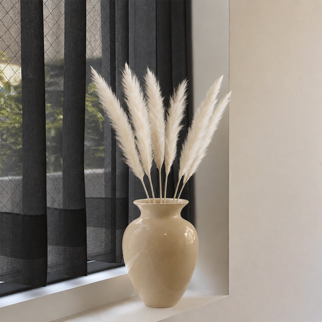 A beige ceramic vase filled with fluffy white pampas grass, placed on a windowsill next to dark gray sheer curtains.