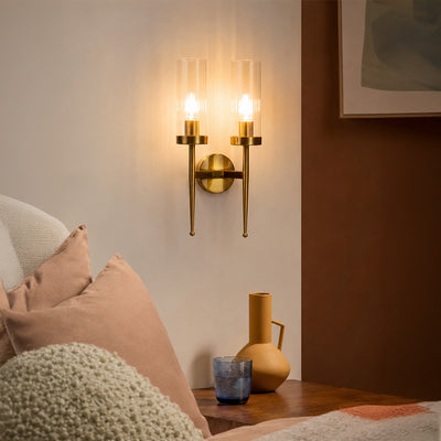 Modern gold brass double candle wall sconce glowing warmly over a cozy bedroom nightstand corner setup with pillows.