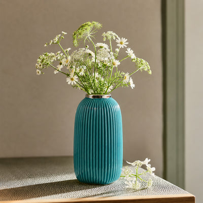 Lifestyle image of a teal fluted ceramic vase styled with white wildflowers on a rustic tabletop with warm sunny shadows.