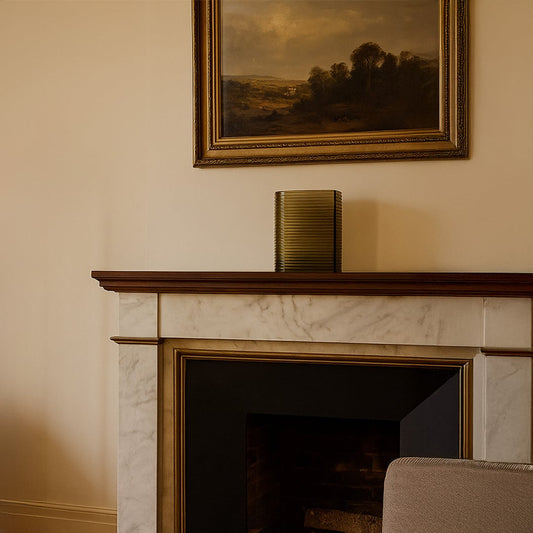 Decorative ribbed metal vase placed on classic fireplace mantel beneath framed artwork creating warm elegant living room decor accent.