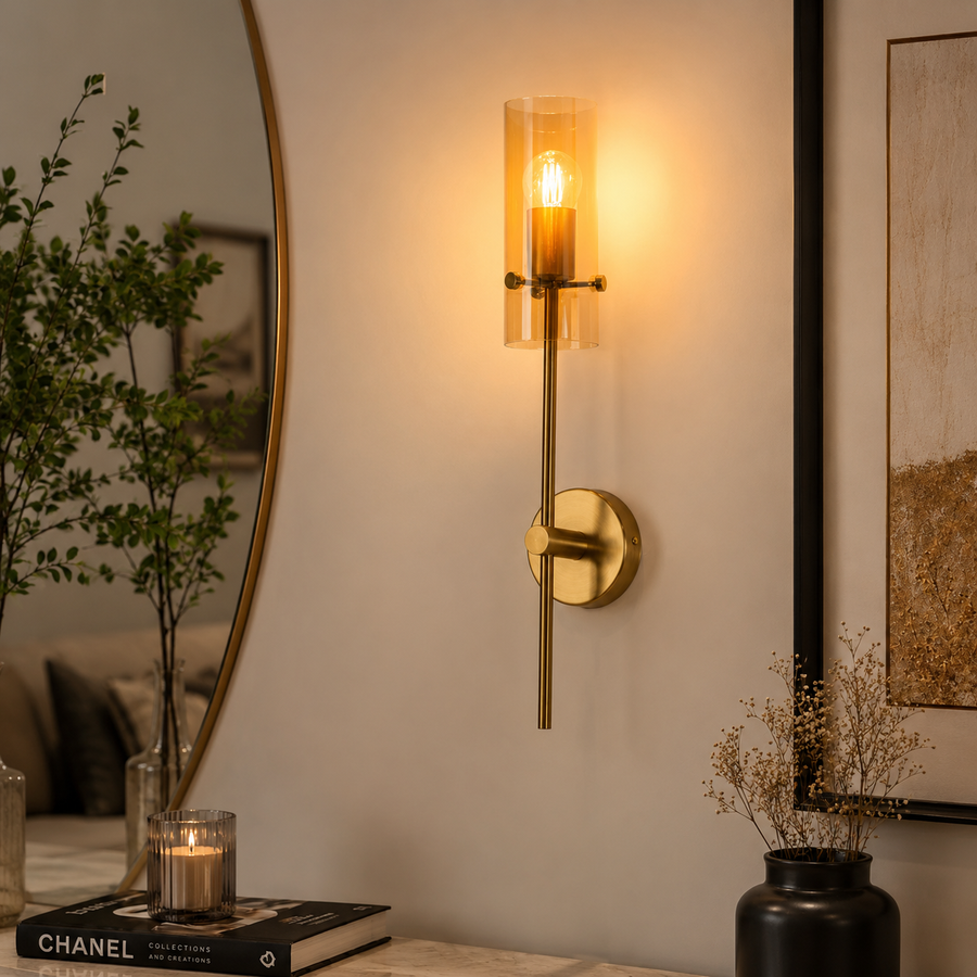 A modern wall sconce with a vertical clear glass cylinder shade and a slim brass-toned metal stem, mounted on a beige wall in a styled interior setting with a mirror, framed art, and dried flowers.