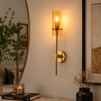 A modern wall sconce with a vertical clear glass cylinder shade and a slim brass-toned metal stem, mounted on a beige wall in a styled interior setting with a mirror, framed art, and dried flowers.