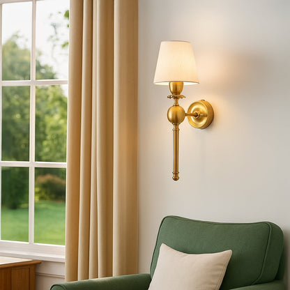 Classic gold torch wall sconce lamp with a cream fabric shade casting warm light over a cozy green living room armchair.