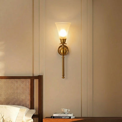 A warm brass wall sconce with a frosted glass shade glowing with a soft yellow light, mounted on a beige paneled wall above a wooden headboard and bedside table.