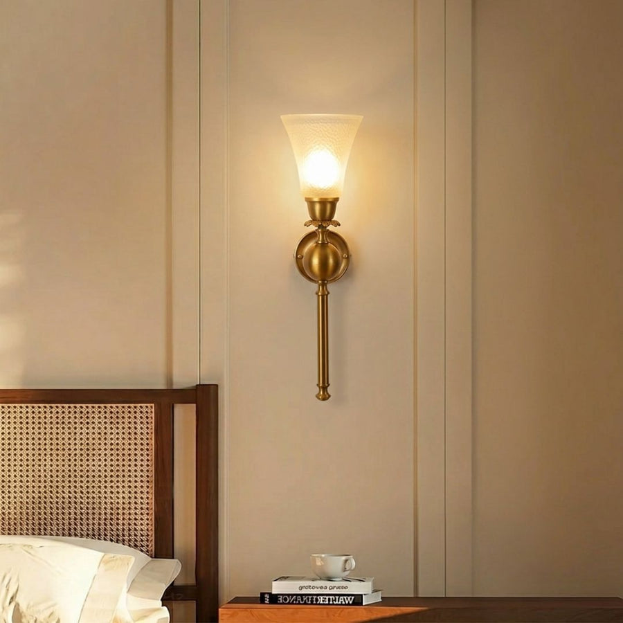 A warm brass wall sconce with a frosted glass shade glowing with a soft yellow light, mounted on a beige paneled wall above a wooden headboard and bedside table.