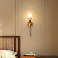 A warm brass wall sconce with a frosted glass shade glowing with a soft yellow light, mounted on a beige paneled wall above a wooden headboard and bedside table.