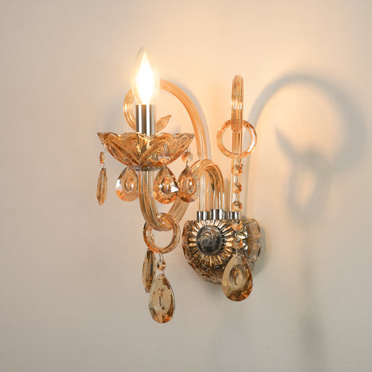 An ornate wall sconce with amber-tinted glass arms and hanging teardrop crystals, shown lit against a plain light wall with a soft shadow cast to the right.