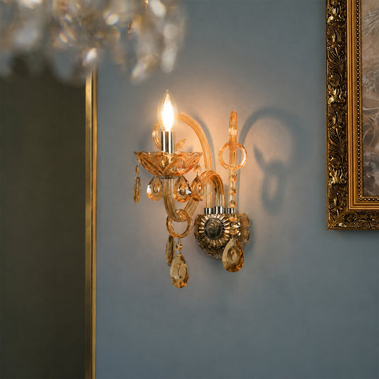 An ornate gold and crystal wall sconce illuminated on a muted blue-gray wall, with part of a gold-framed artwork and a hanging chandelier visible in the scene.