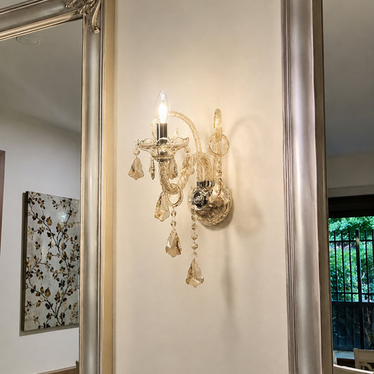 A vintage-style crystal and gold wall sconce mounted on a beige wall, flanked by ornate silver-framed mirrors reflecting a home interior.