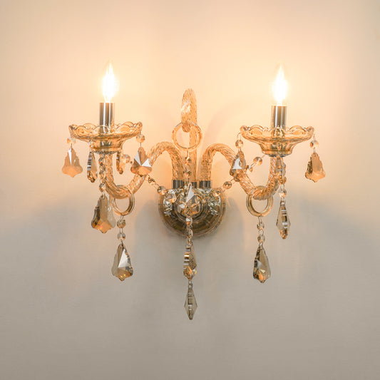 A two-light wall sconce with a warm gold-toned metal frame, ornate curved arms, and hanging clear crystal teardrop pendants, shown lit against a plain light wall.