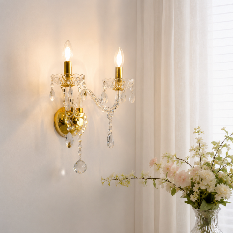 A two-light wall sconce with a polished gold finish and clear crystal droplets, mounted on a white wall next to a window with sheer curtains and a vase of white flowers in the foreground.